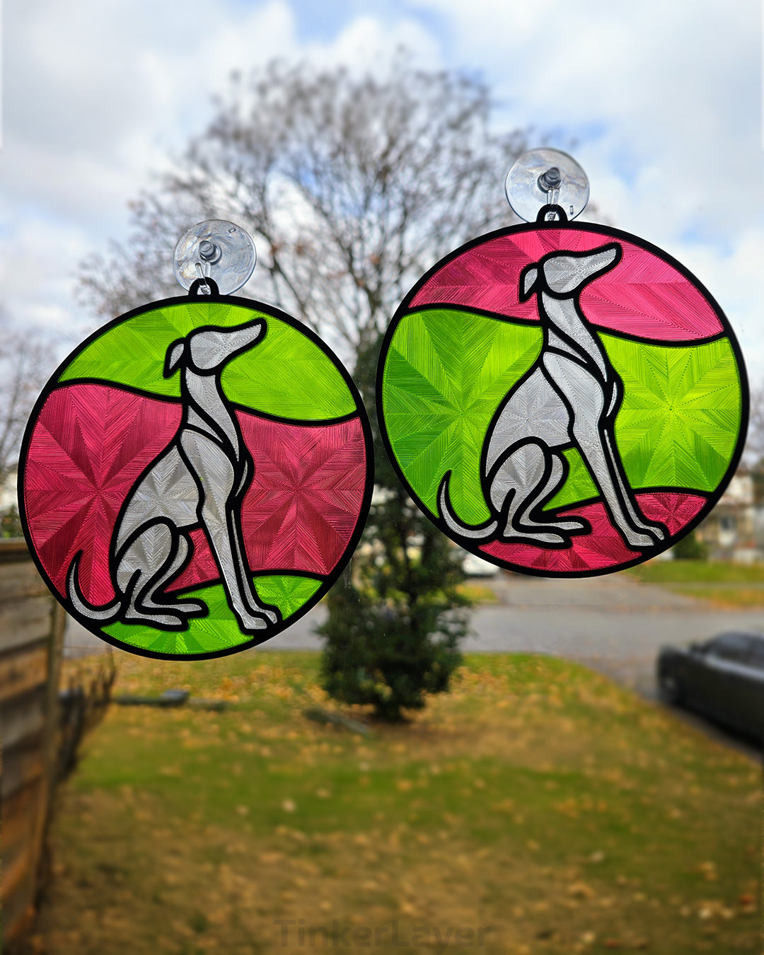 Two round whippet stained glass style suncatchers with red and green accents hanging in a window